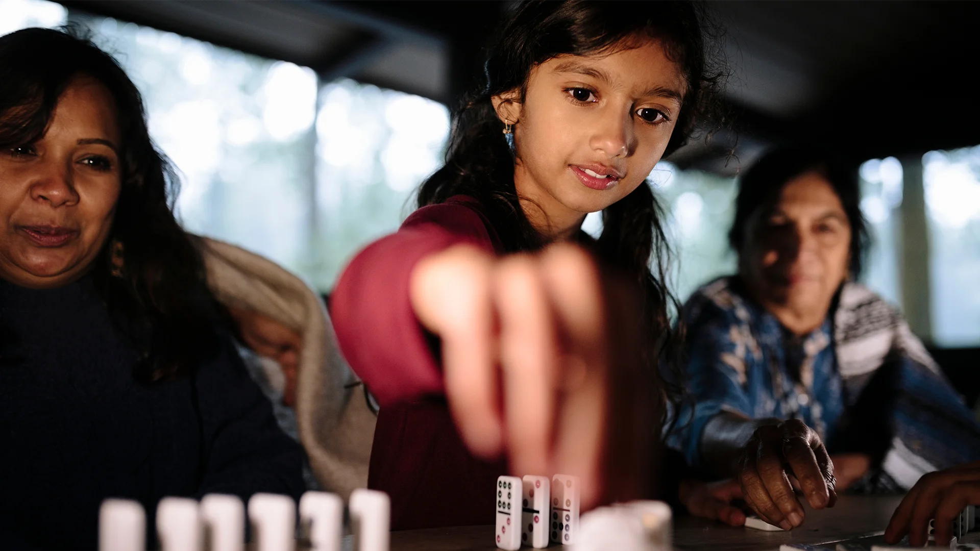 Young girl playing domiinoes next to two women (photo)