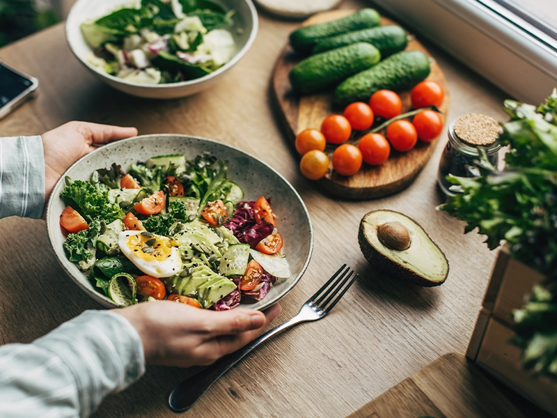 Hands holding a bowl of fresh salad with avocado, tomatoes, and egg on a wooden table. (photo)