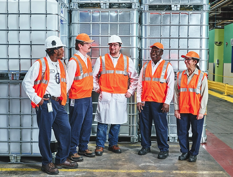 Five factory workers in safety gear standing together in front of large storage containers. (photo)