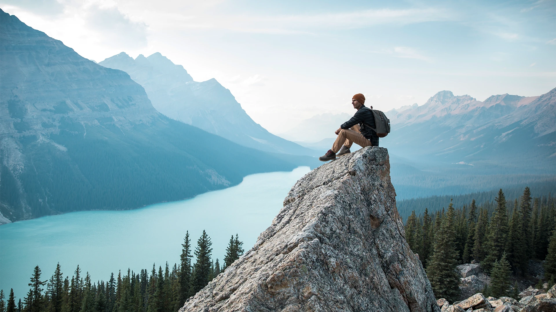 A hiker sits on a rocky cliff overlooking a turquoise mountain lake, surrounded by forested slopes and distant peaks. (photo)