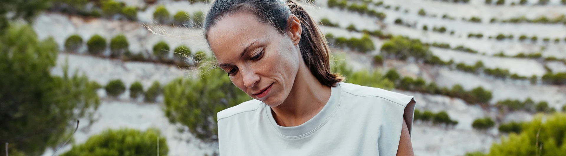 A woman looks down thoughtfully outdoors with terraced greenery in the background. (photo)