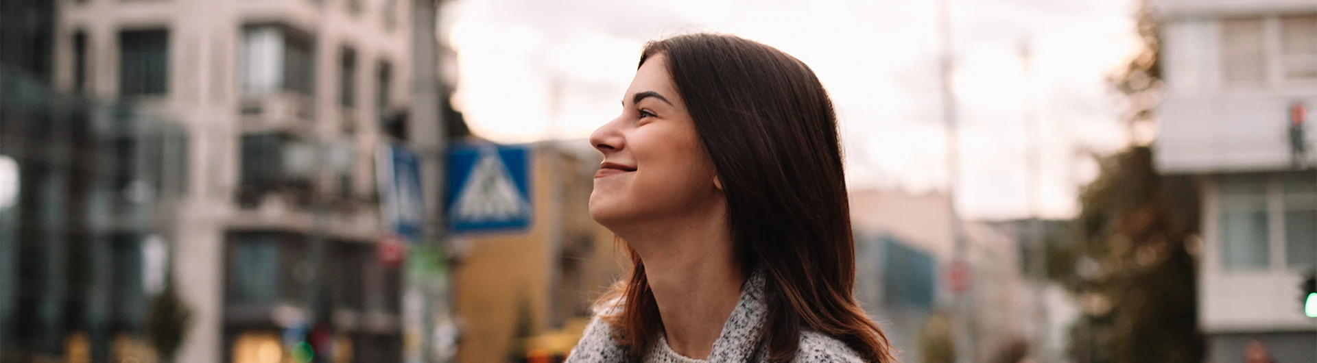 A woman smiles gently while looking ahead on a city street, with urban buildings softly blurred in the background. (photo)