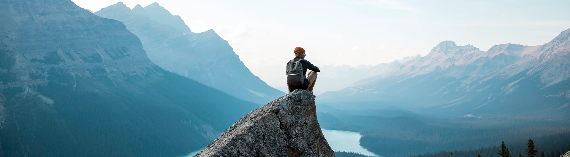 A hiker with a backpack sits on a rocky peak overlooking a vast mountain landscape and valley below. (photo)
