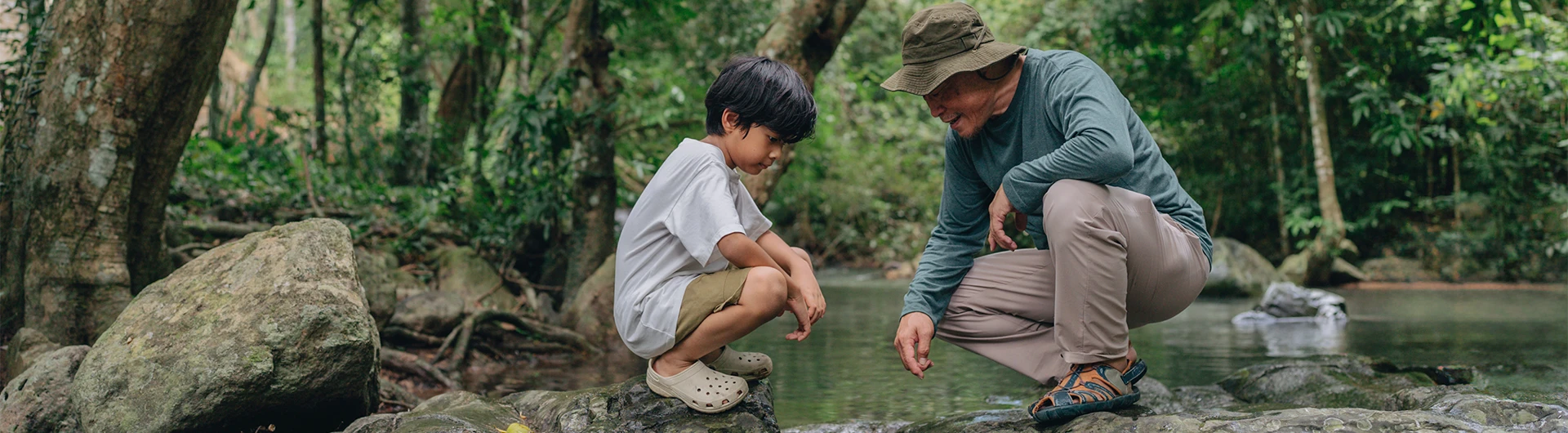 A man and a young boy crouch by a forest stream, observing something in the water together. (photo)