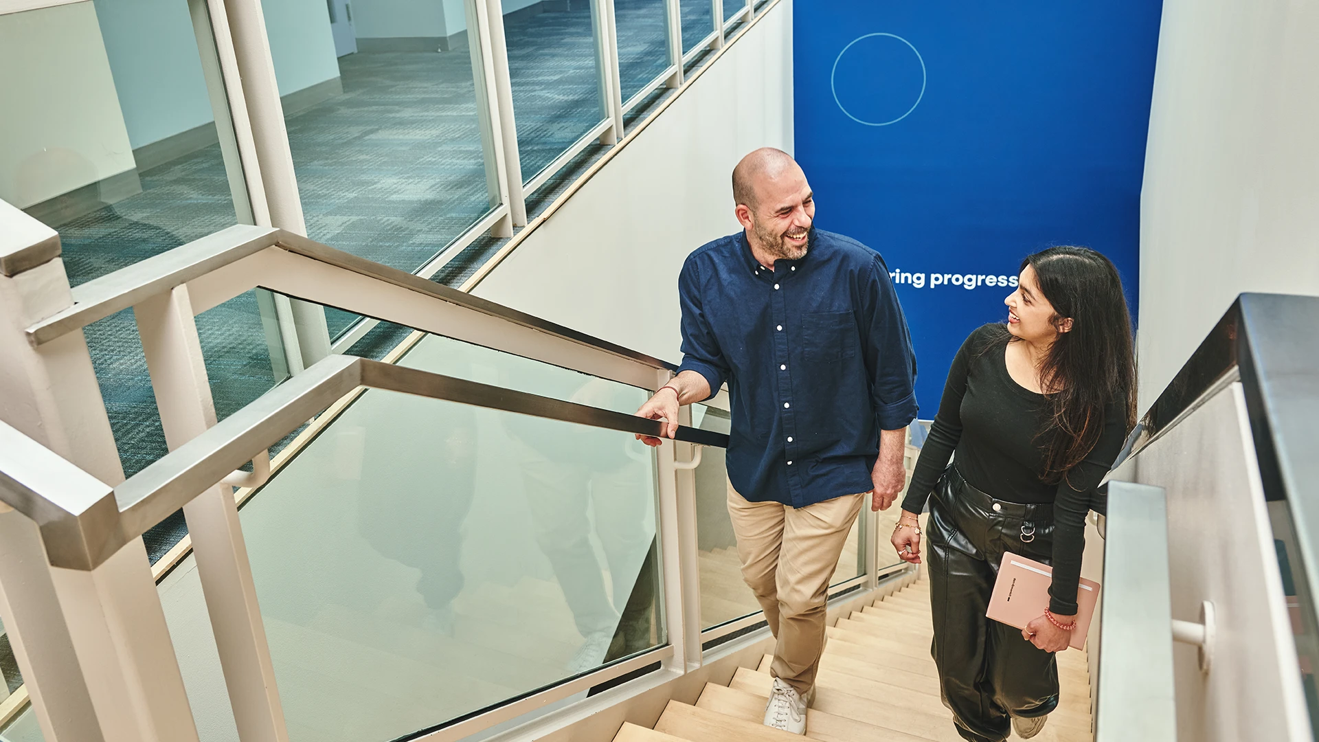 Two colleagues walk up a modern office staircase, smiling and talking while holding notebooks. (photo)