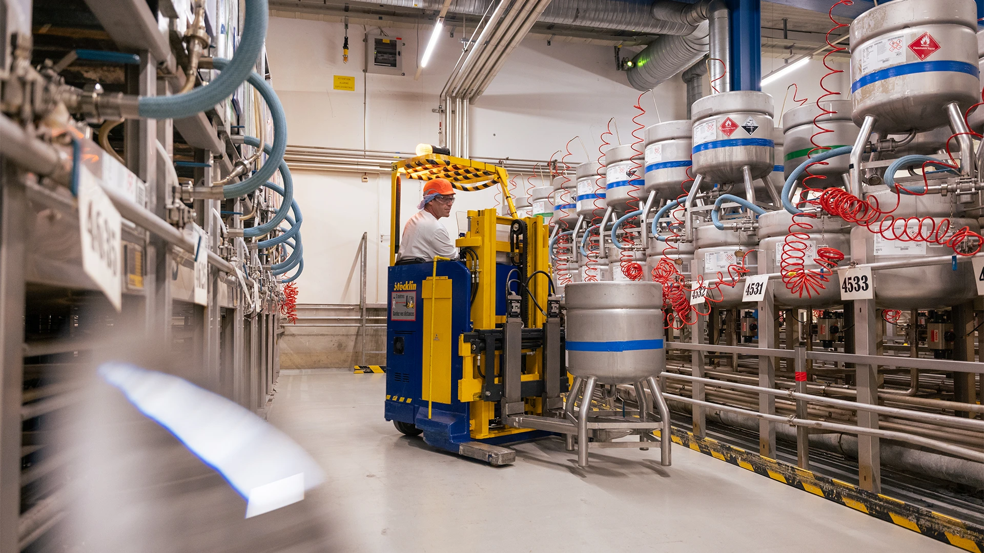 An industrial facility interior where a worker operates a specialized forklift among rows of large stainless steel containers connected by hoses and piping, in a clean, well-lit production environment. (photo)