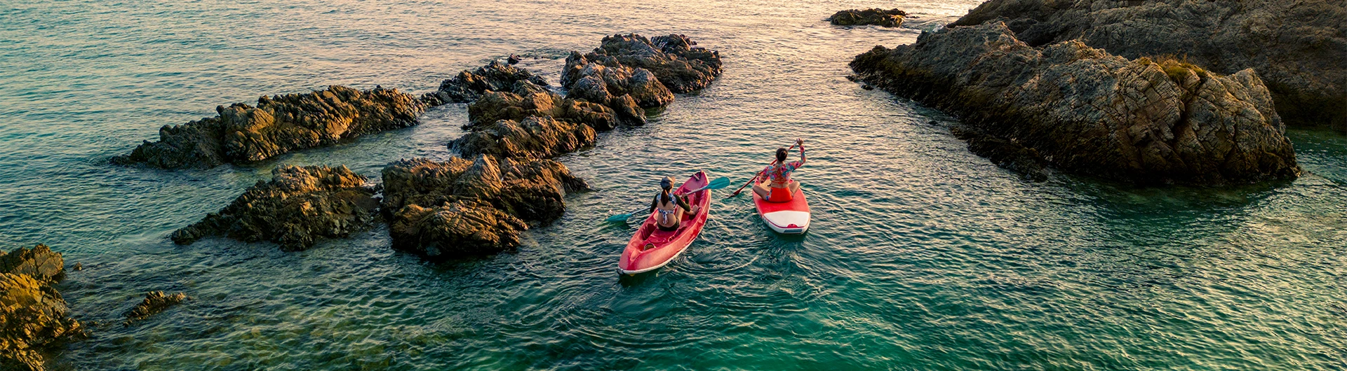 Two people paddle kayaks through calm turquoise water between rocky outcrops at sunset. (photo)