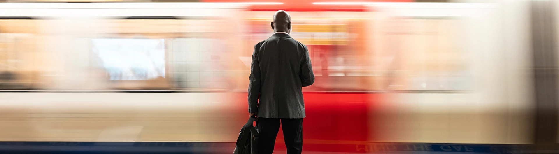 A man standing on a train platform, seen from behind, as a train passes by in motion blur. (photo)