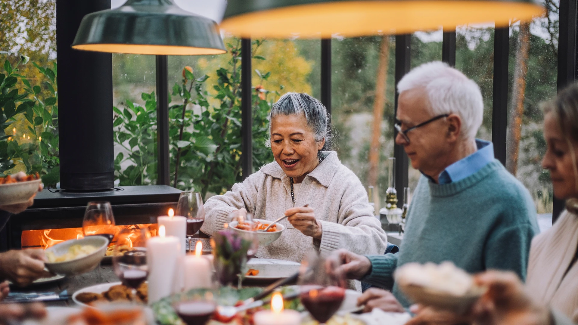 An elderly woman serves food at a warmly lit dinner table while others share a meal together indoors. (photo)