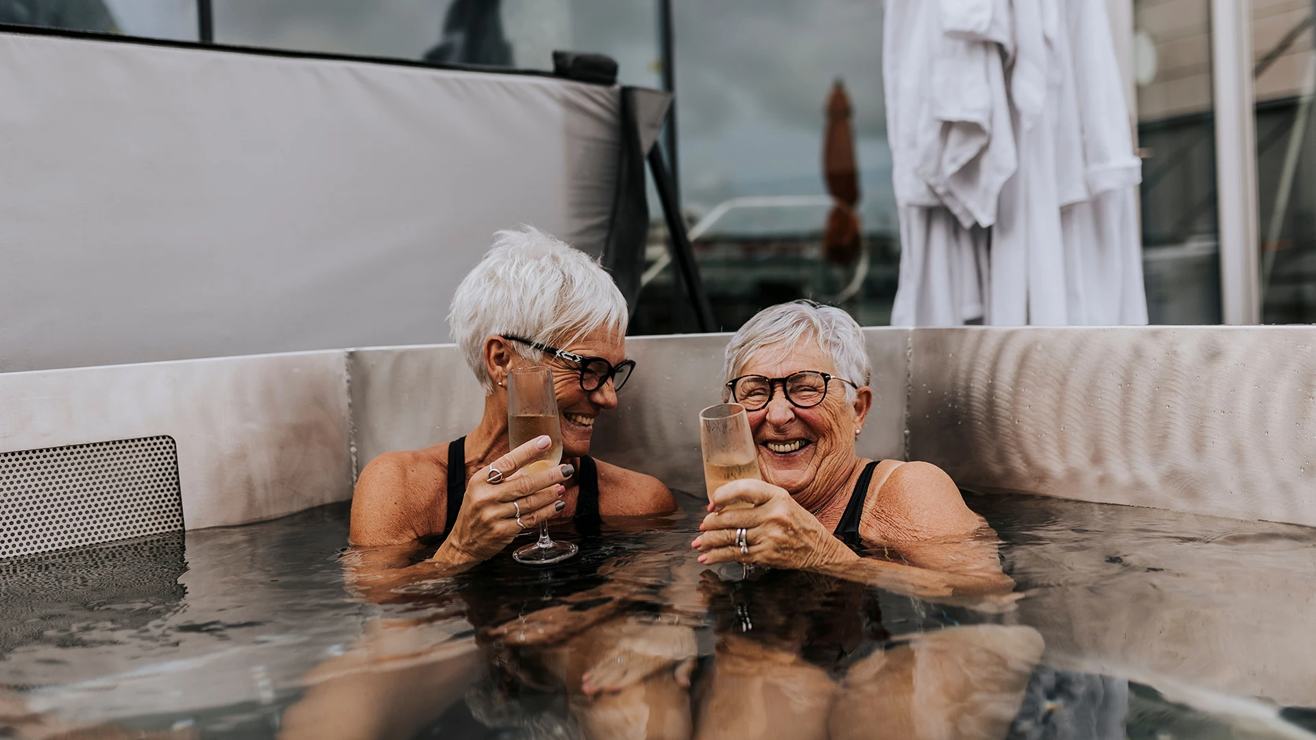 Two elderly women smile and clink glasses while relaxing together in an outdoor hot tub. (photo)