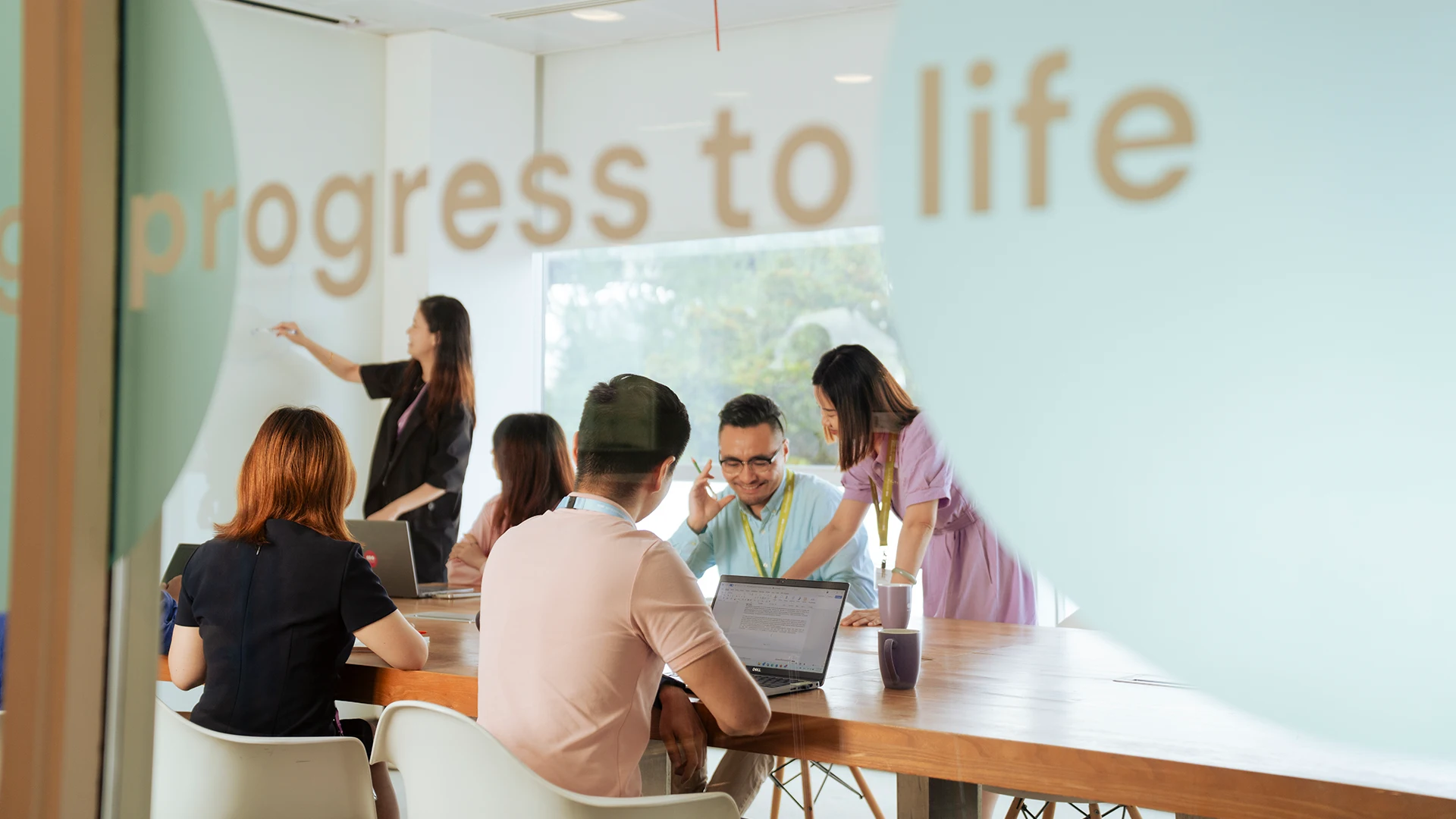 A diverse team collaborates around a table in a bright meeting room with “progress to life” written on the glass. (photo)