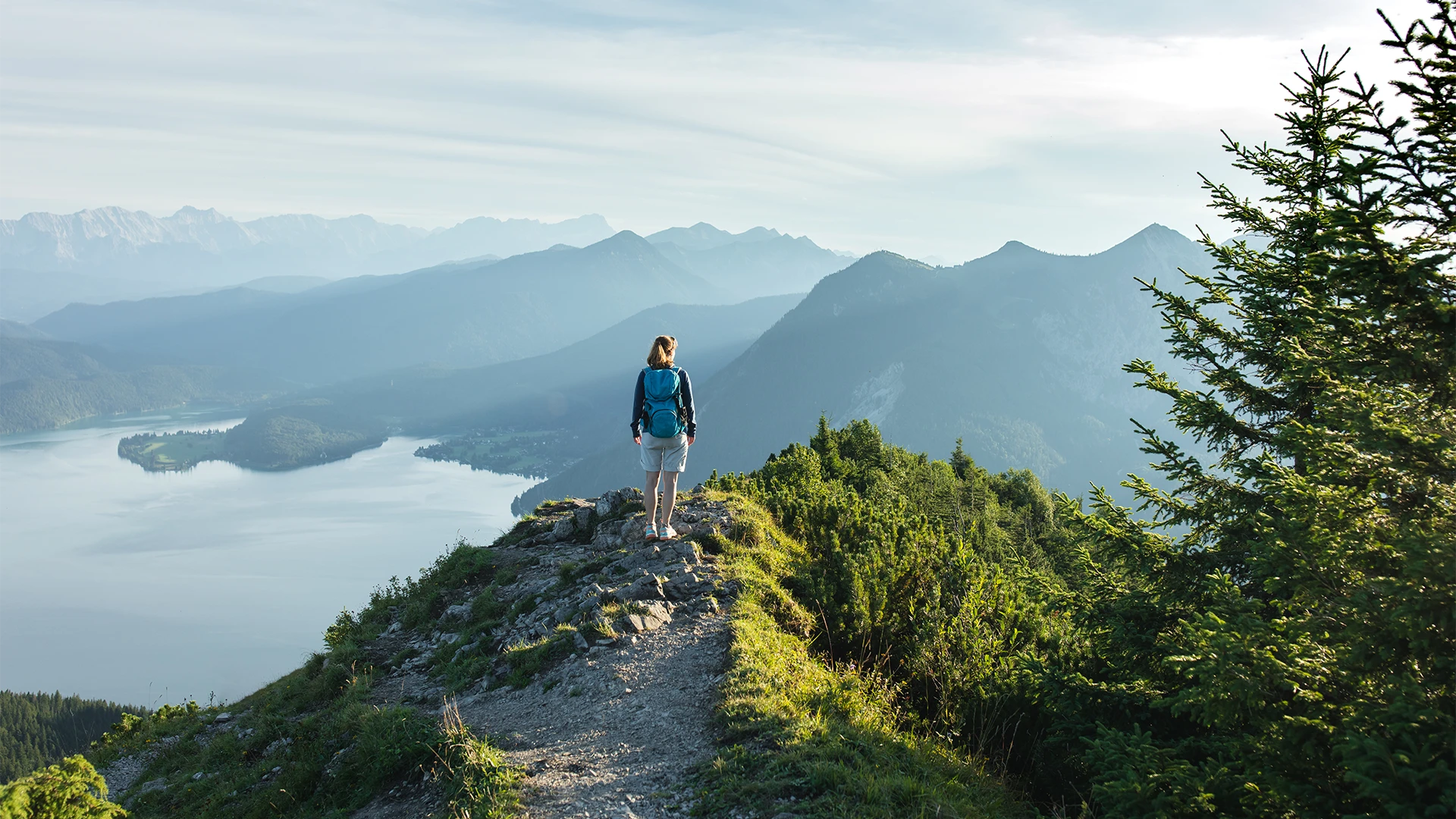 A hiker stands on a narrow mountain ridge overlooking a lake and layered alpine peaks in the distance. (photo)
