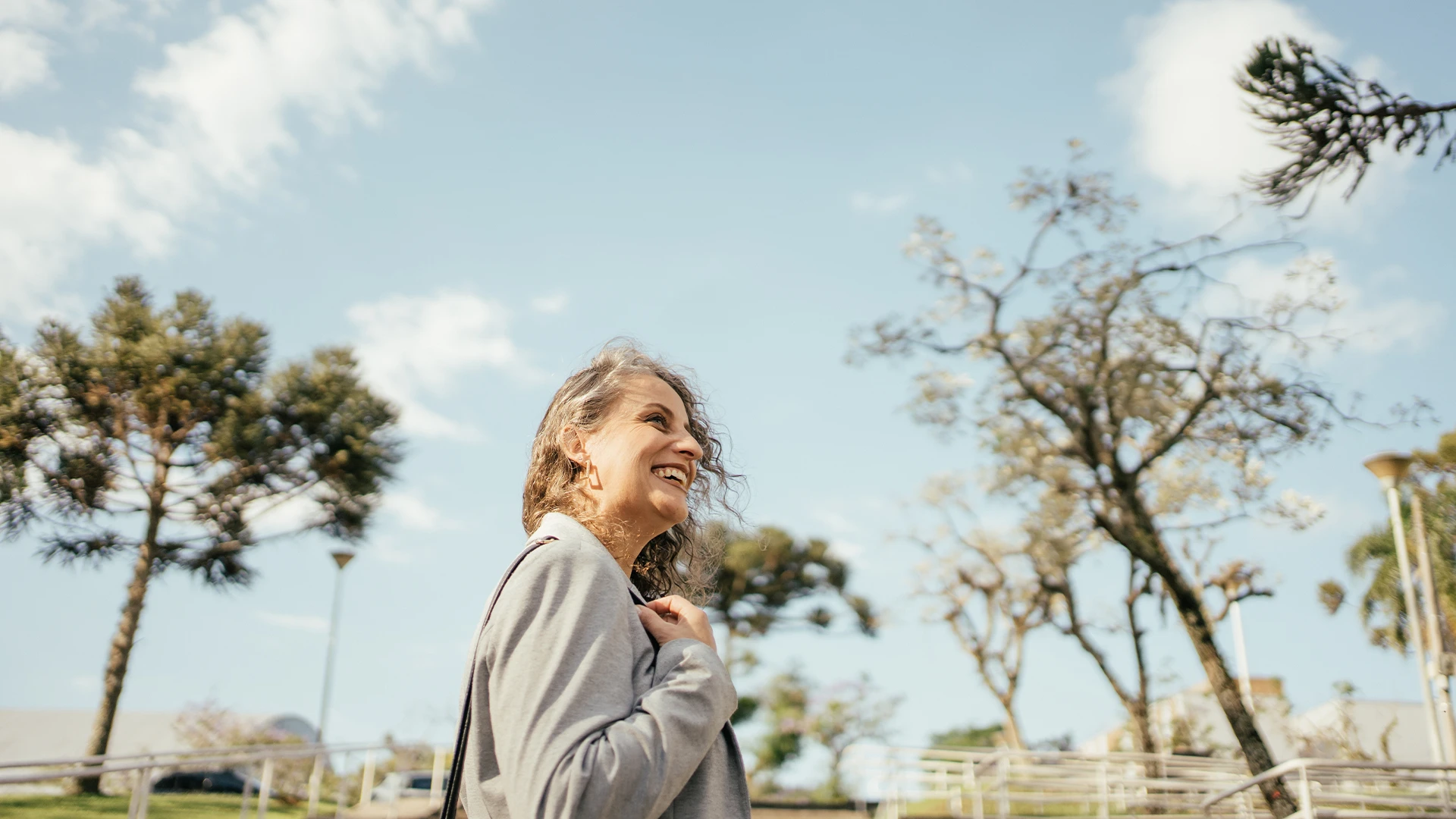 A woman smiles while walking outdoors through a park-like setting with trees under a bright sky. (photo)