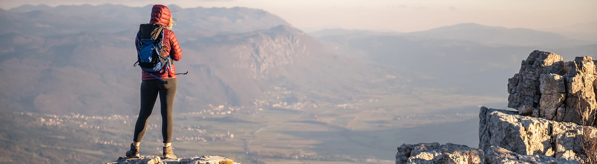A person wearing a backpack stands on a rocky viewpoint, looking out over a wide mountain landscape. (photo)