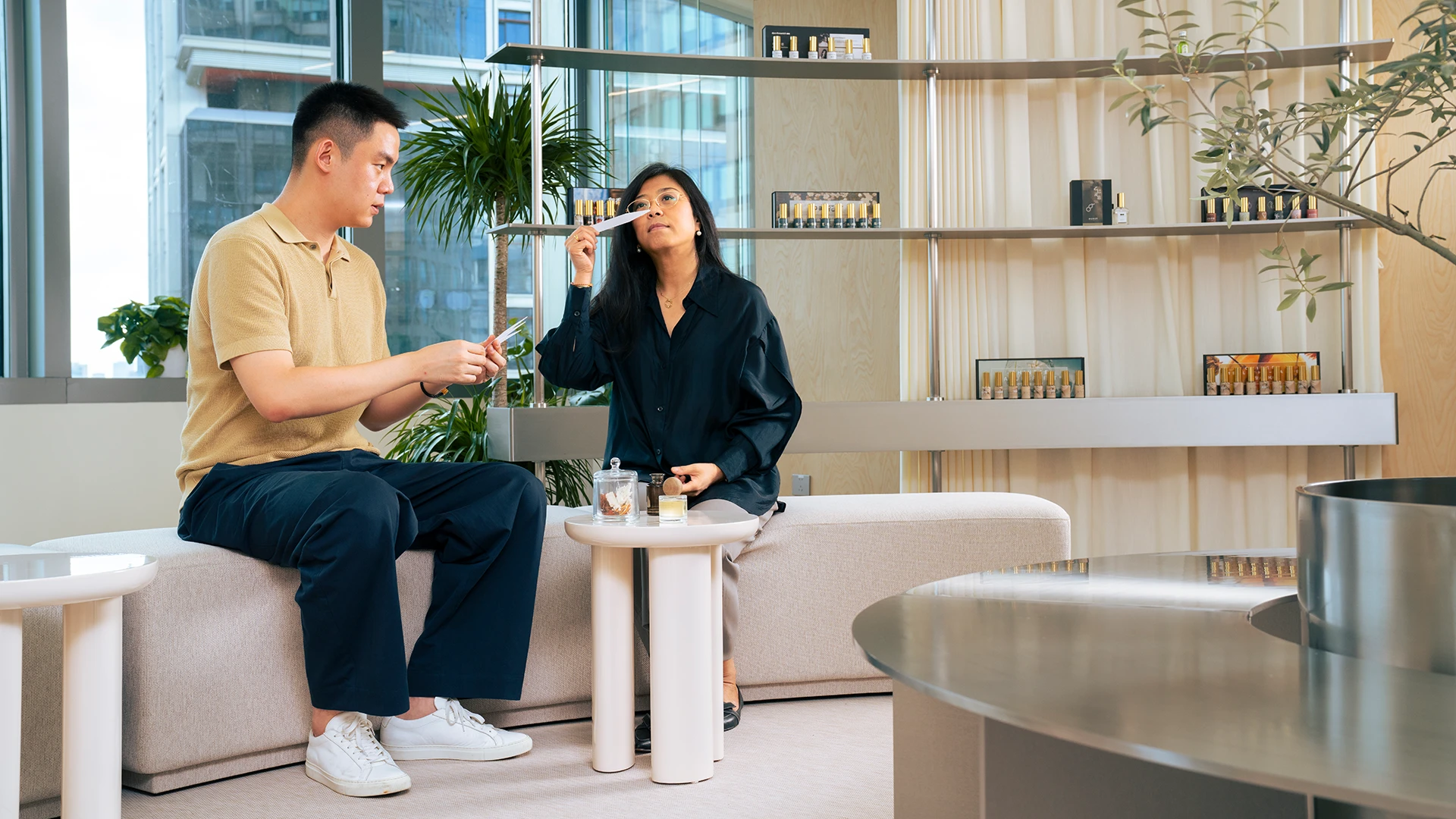 Two people sit in a modern showroom, smelling perfume samples and discussing fragrances surrounded by displayed bottles. (photo)