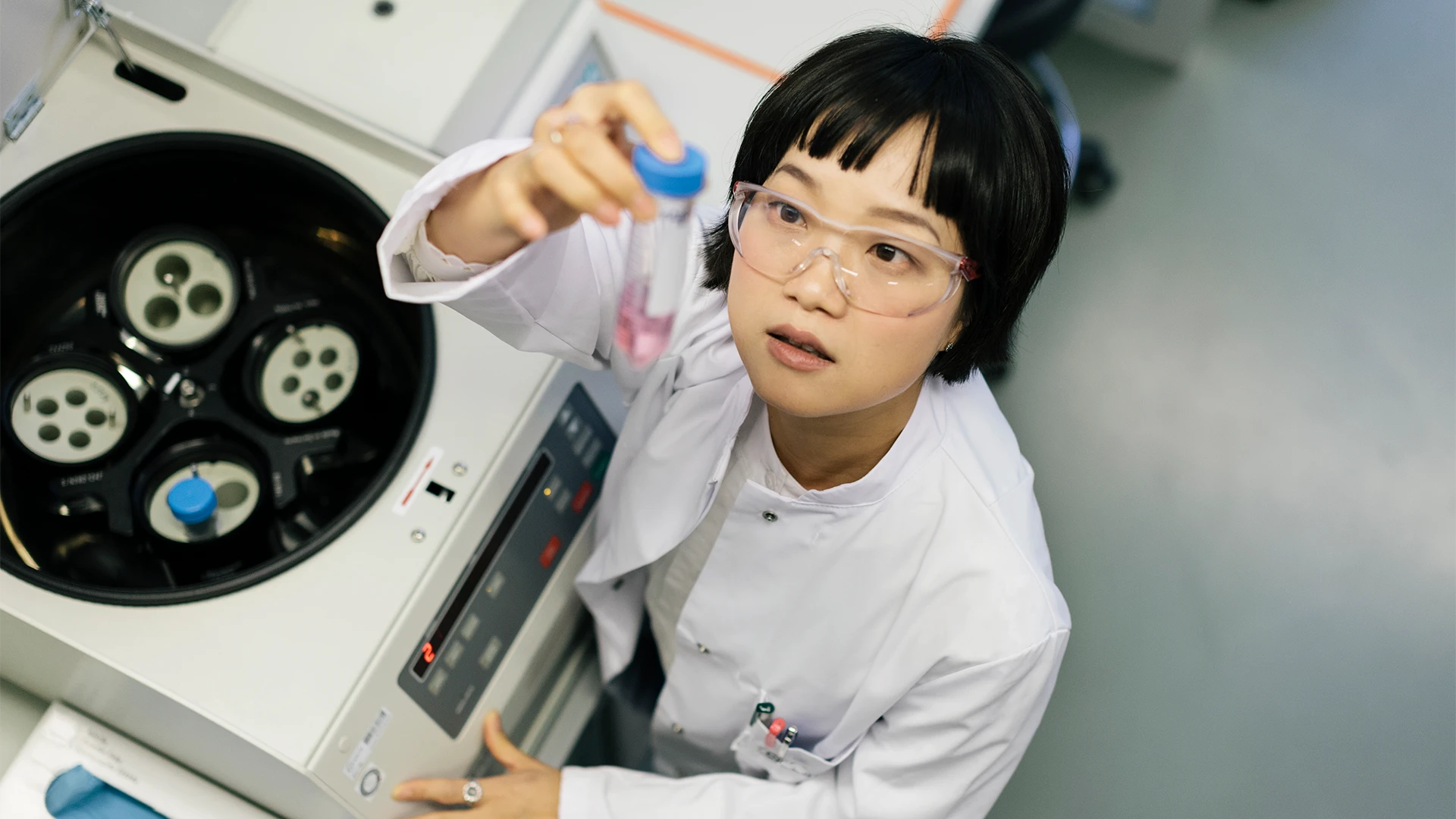 A scientist in a lab coat and safety goggles examines a small sample tube, holding it up beside a laboratory centrifuge in a clean, modern research environment. (photo)