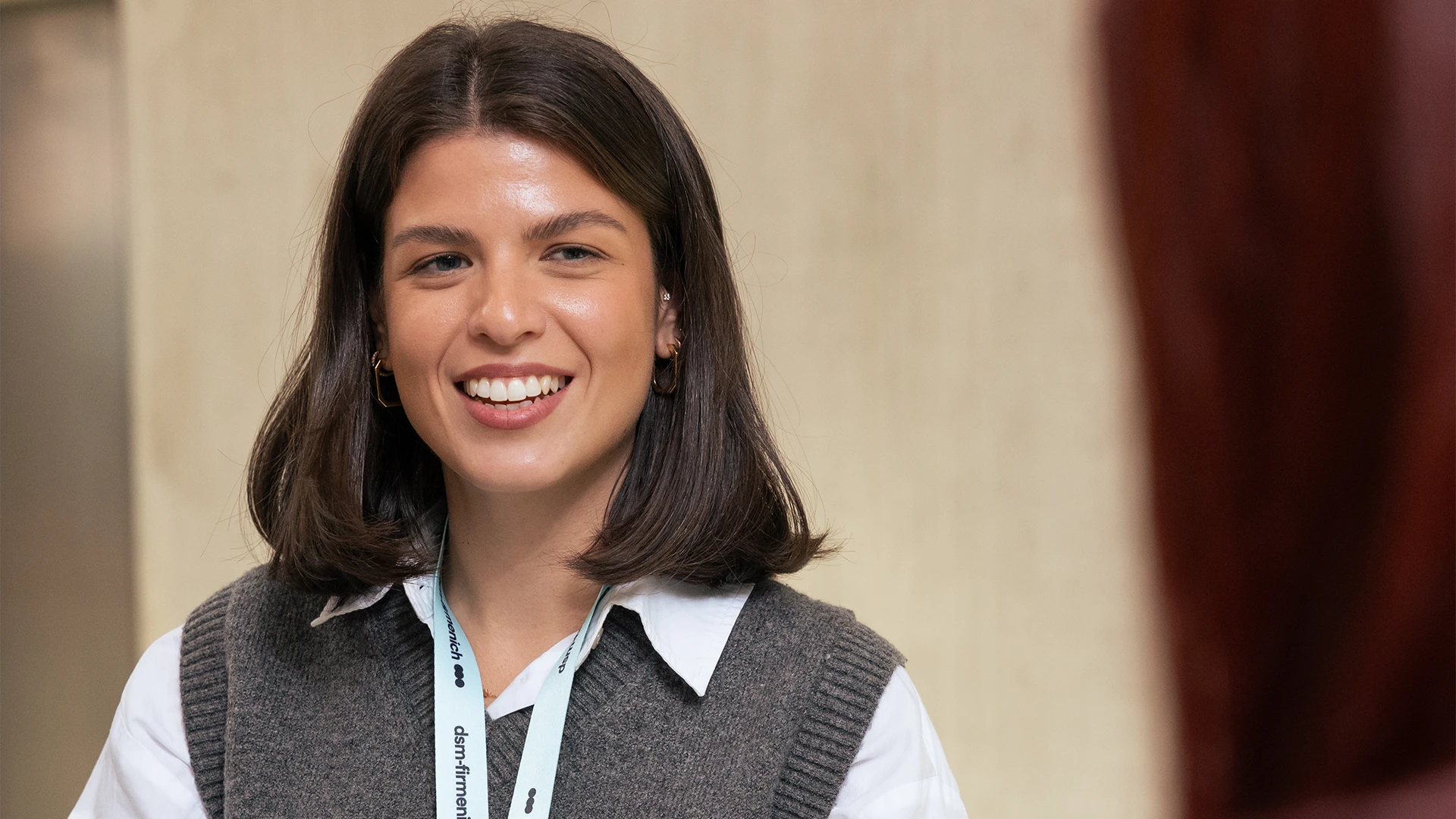A young professional smiles warmly in an office setting, wearing a lanyard and business-casual attire, with a softly blurred background suggesting a conversation or meeting. (photo)