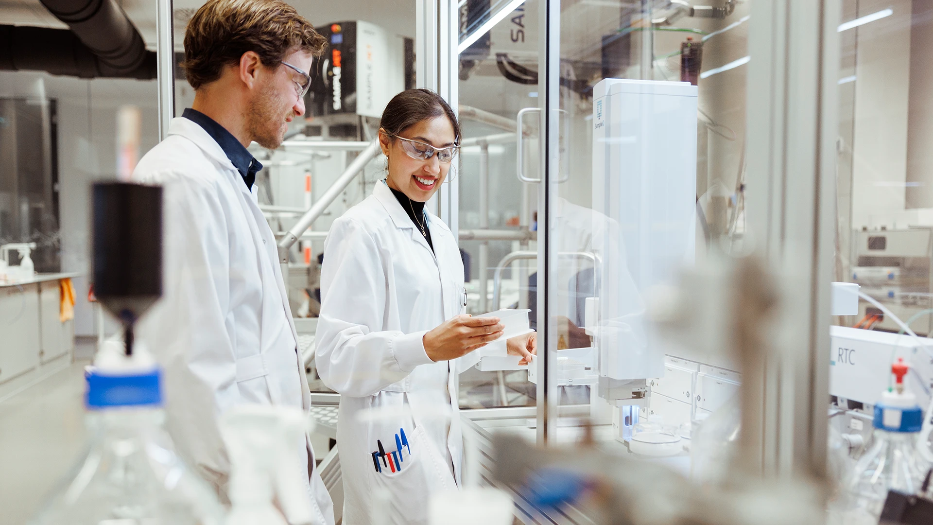 Two researchers in lab coats review samples together while working with automated equipment in a laboratory. (photo)