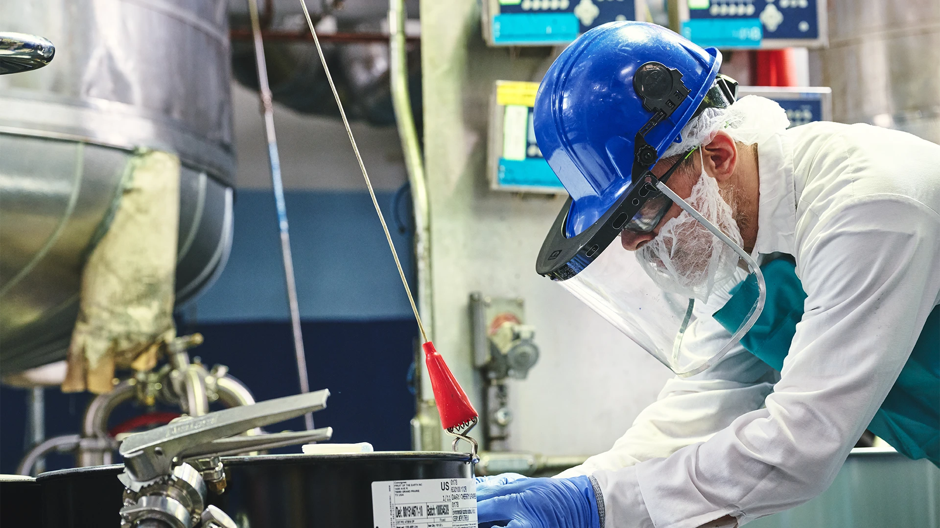 A factory worker wearing a hard hat, face shield, hair net, and gloves carefully inspects equipment on an industrial production line. (photo)