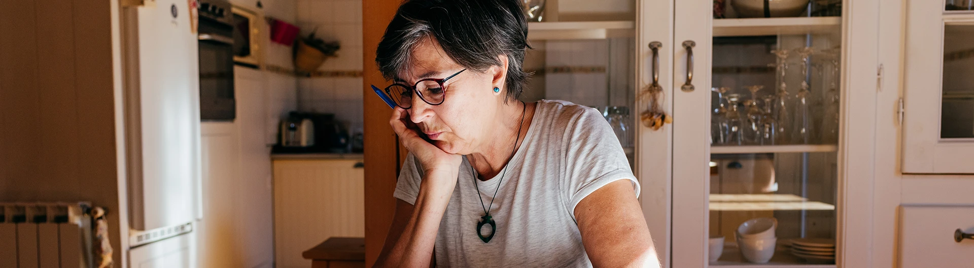 A woman wearing glasses rests her head in her hand while sitting thoughtfully at a kitchen table. (photo)