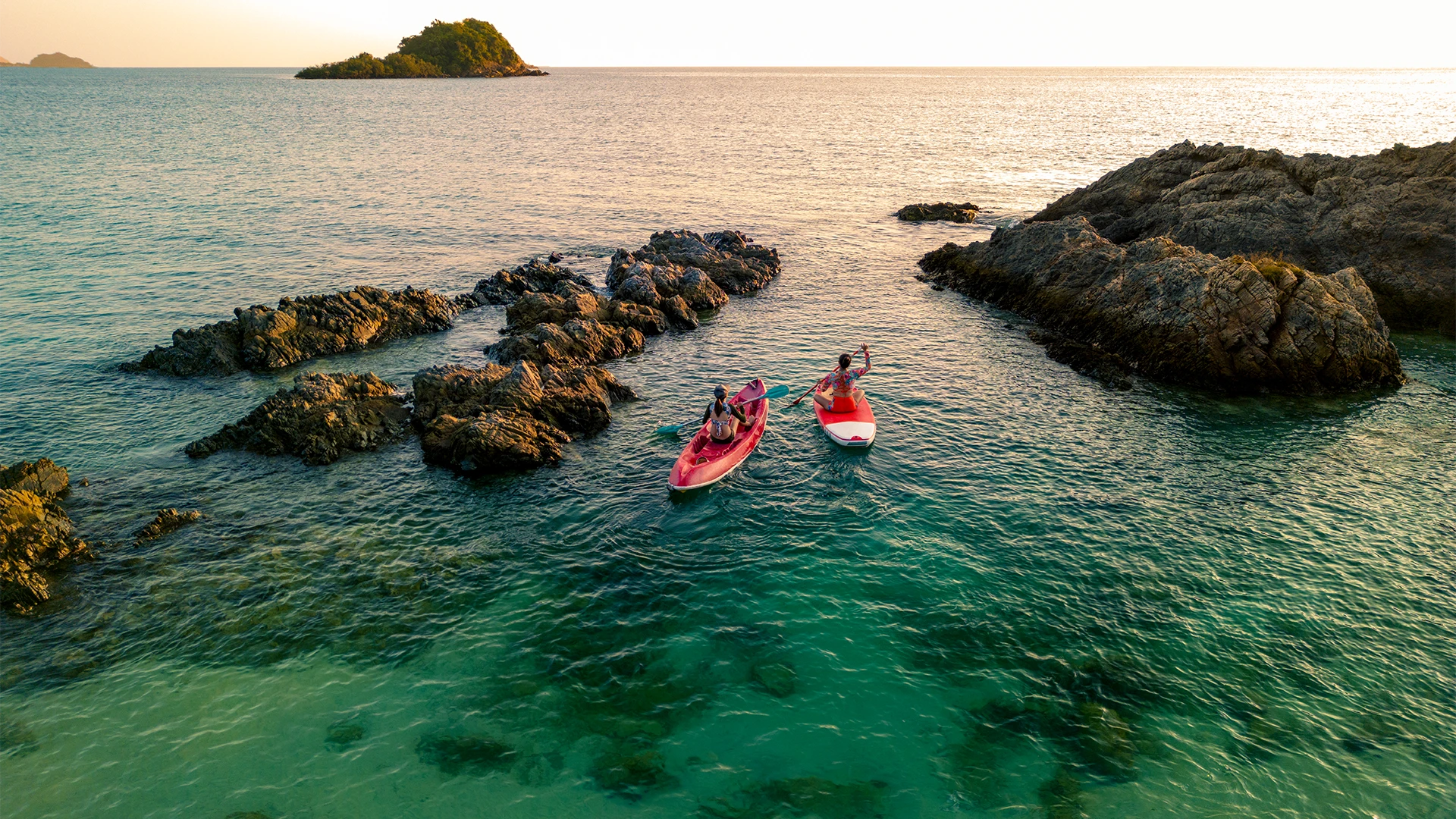 Two people paddle a kayak and a paddleboard through clear turquoise water among rocky islets at sunset. (photo)