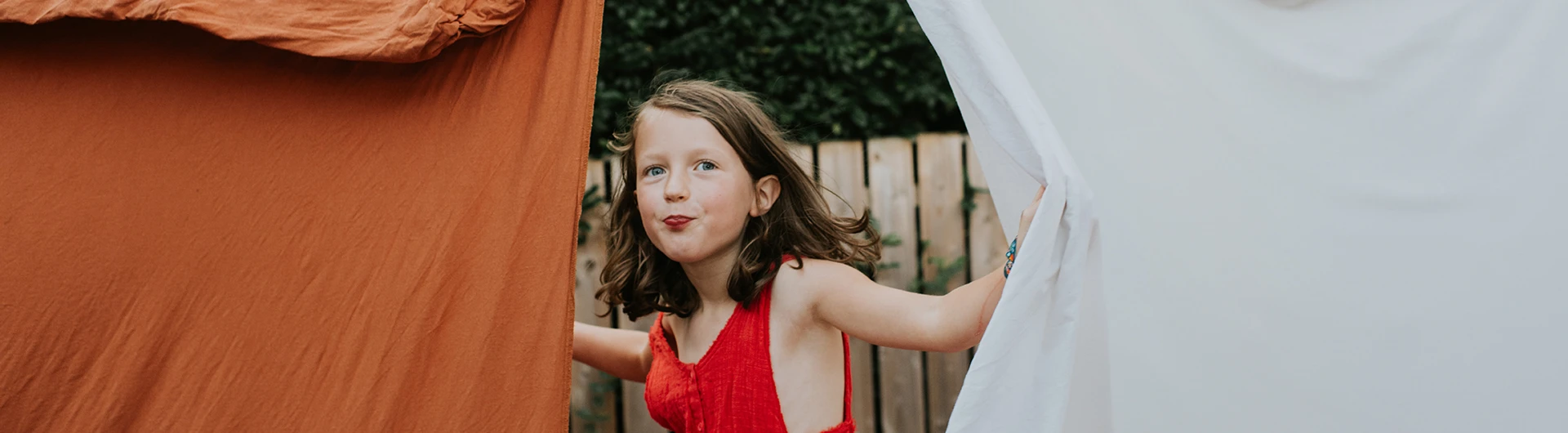 A child peeks out between hanging fabric sheets outdoors. (photo)