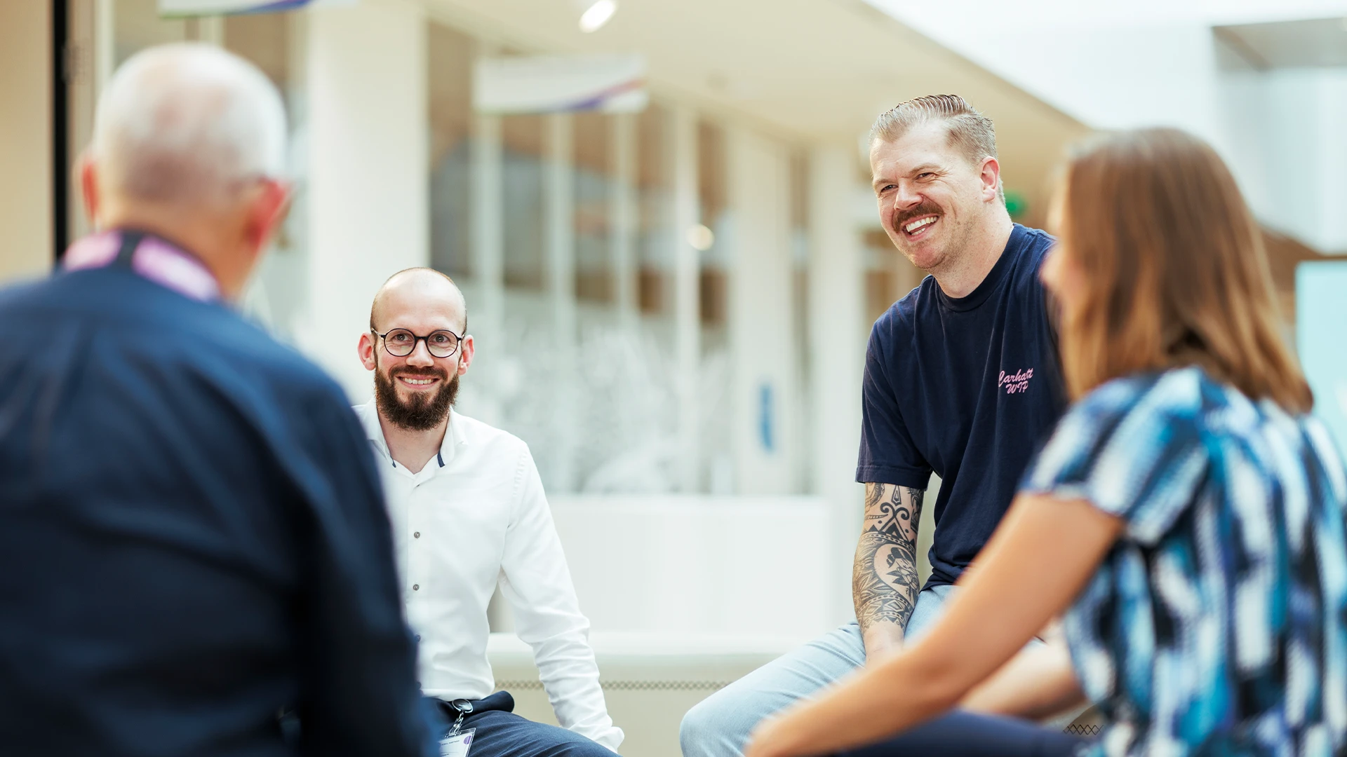 A group of colleagues sit together indoors, smiling and chatting in a relaxed workplace setting. (photo)