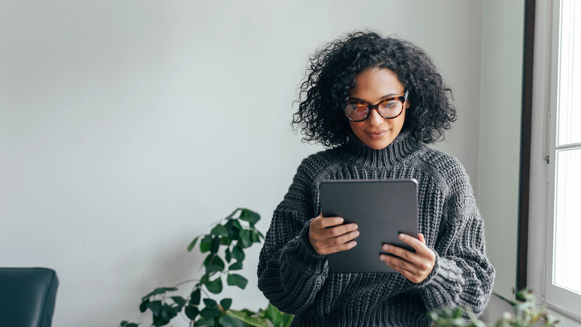 A woman with curly hair and glasses stands in a bright, minimalist room, calmly reading from a tablet, with soft daylight and a houseplant in the background. (photo)