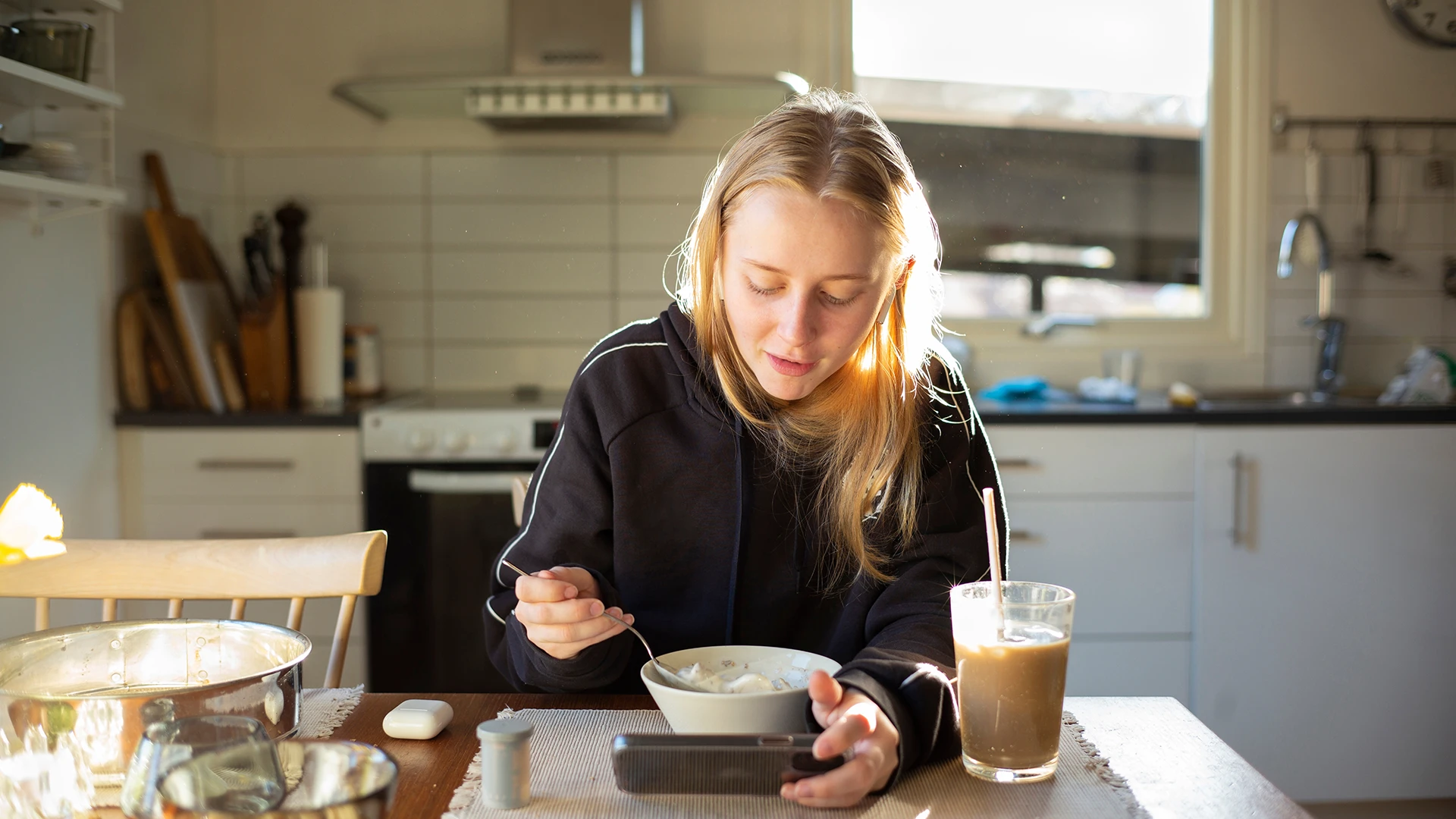 A young woman eats breakfast at a kitchen table while looking at her phone in soft morning light. (photo)