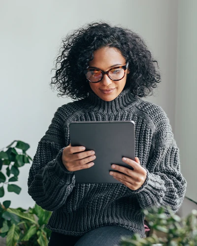 A woman with glasses reads content on a tablet while standing indoors near green plants. (photo)