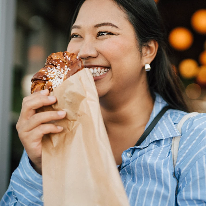 A smiling woman holds a pastry in a paper bag while enjoying a bite outdoors. (photo)