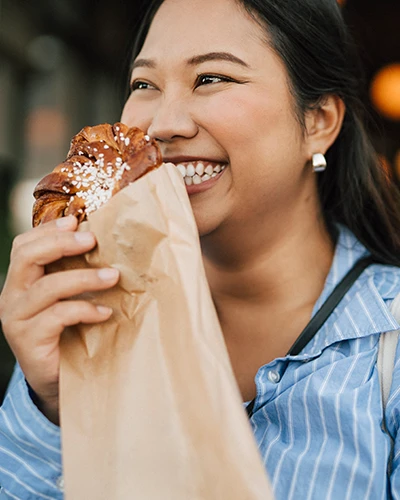 A smiling woman holds a pastry in a paper bag while enjoying a bite outdoors. (photo)