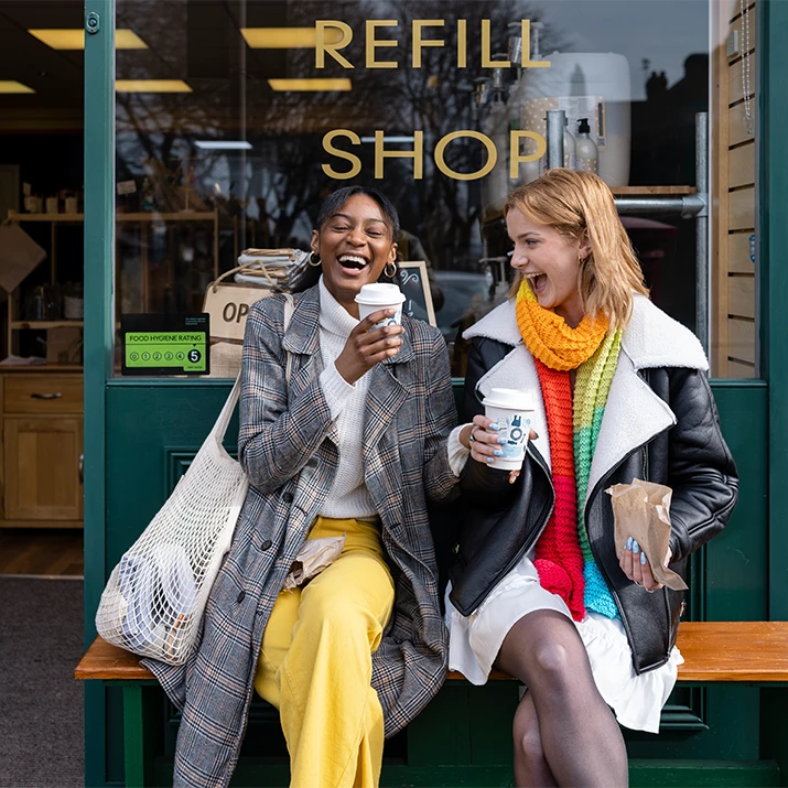 Two women sitting outside a shop, laughing and holding cups. (photo)