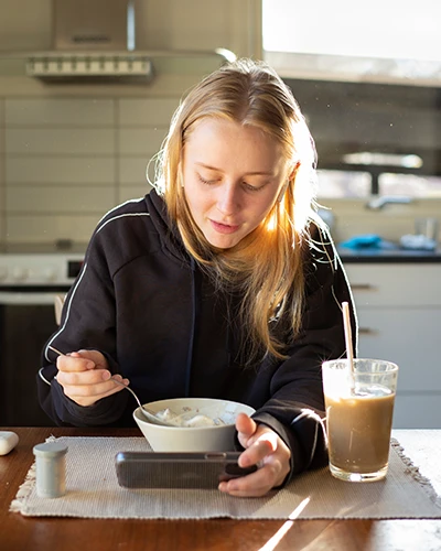Young woman eating breakfast while looking at her phone. (photo)
